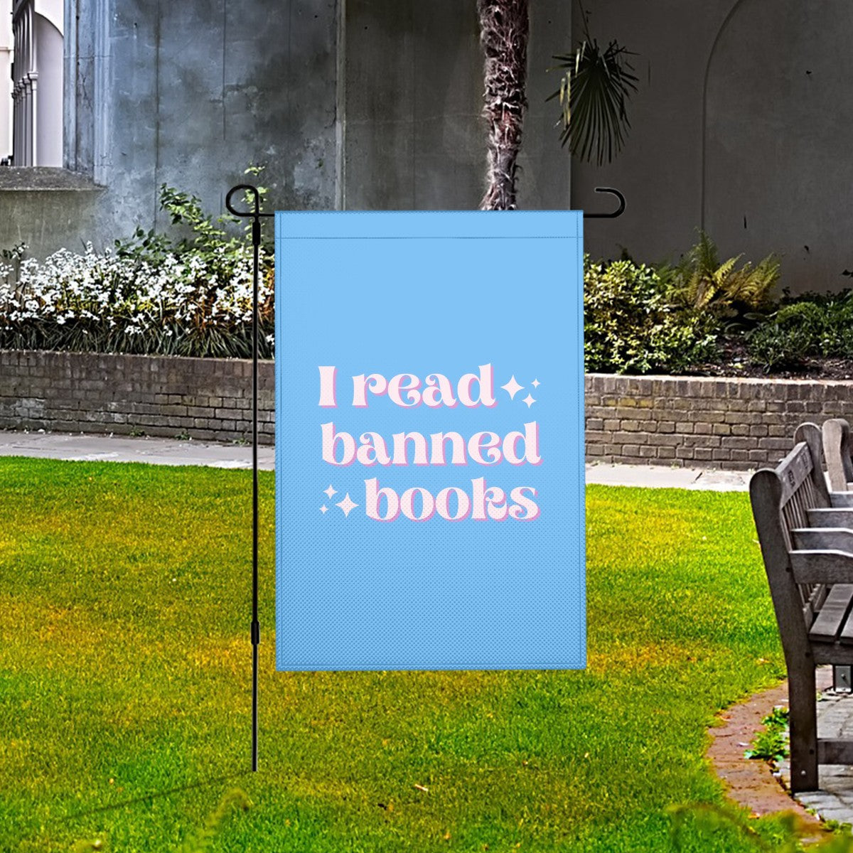 Garden flag with 'I read banned books' text on a grassy area with a bench and plants.